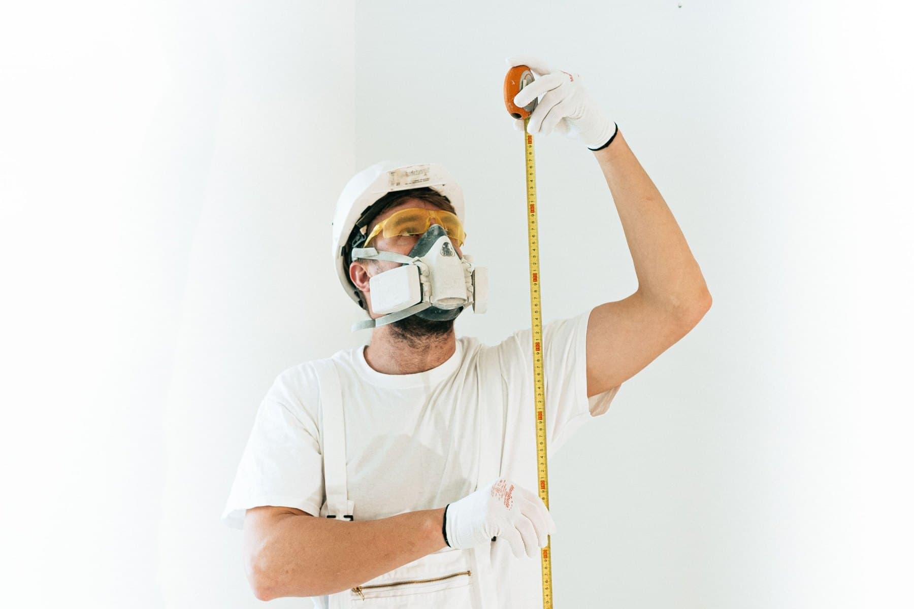 Un artisan en tenue blanche, casque et masque, mesure une hauteur avec un mètre ruban